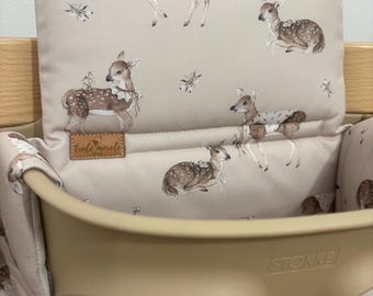 May include: A beige high chair cushion featuring a repeating pattern of brown and white fawns with small floral accents. The cushion includes a rectangular brown leather-like tag with the text trele morele shop and fits into a beige Stokke high chair.