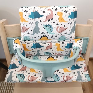 May include: A light blue high chair cushion with a white background and a dinosaur pattern. The cushion has a light blue strap with the word "Stokke" printed on it.