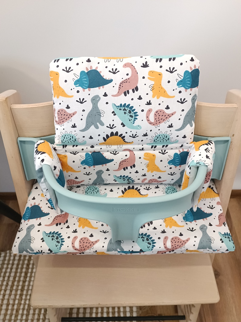 May include: A light blue high chair cushion with a white background and a dinosaur pattern. The cushion has a light blue strap with the word "Stokke" printed on it.