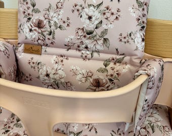 May include: A baby high chair with a pink floral patterned cushion and backrest. The cushion features a repeating pattern of white and brown flowers on a pink background. The high chair is made of light pink plastic and wood.