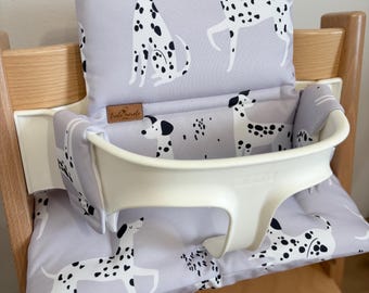 May include: A light gray high chair cushion set featuring a repeating pattern of black and white dalmatian dogs. The padded fabric covers the backrest and seat of a wooden high chair, which includes a cream-colored plastic safety harness attachment.