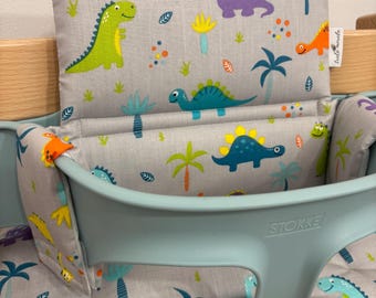 May include: A high chair cushion set with a light blue frame and a gray fabric cover. The fabric features colorful dinosaur illustrations, palm trees, and other tropical elements. The cushion set is designed for a Stokke high chair.