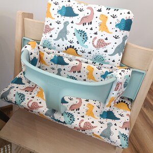 May include: A light blue high chair with a white and multi-coloured dinosaur print cushion. The cushion has a separate headrest and a strap to secure the child in the chair. The chair is made of wood and has a light wood finish.