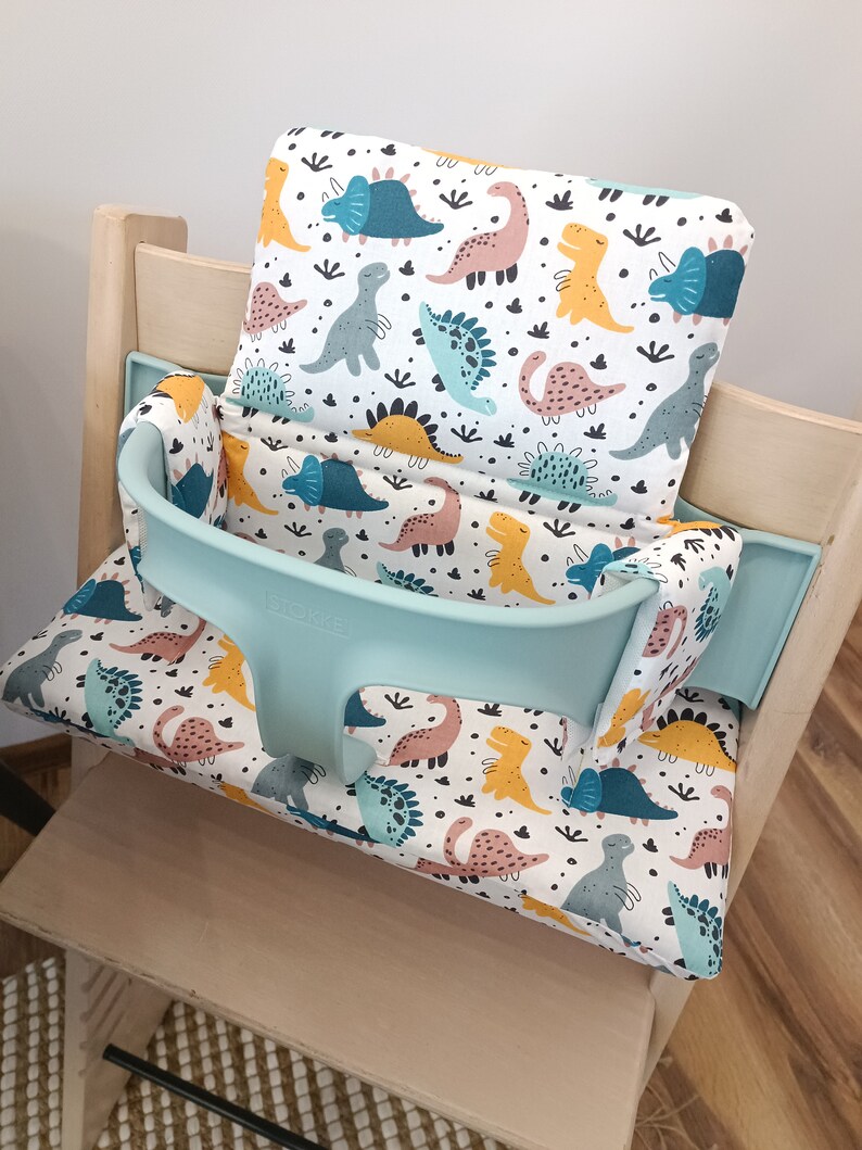 May include: A light blue high chair with a white and multi-coloured dinosaur print cushion. The cushion has a separate headrest and a strap to secure the child in the chair. The chair is made of wood and has a light wood finish.