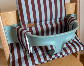May include: A high chair cushion set featuring a vertical striped pattern in light blue and dark red. The fabric covers the backrest and seat of a wooden high chair, which includes a light blue plastic safety rail with the embossed text STOKKE.