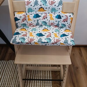 May include: A wooden high chair with a dinosaur-themed cushion set. The cushion set includes a back cushion, a seat cushion, and two side cushions. The cushions are white with a colourful dinosaur pattern.