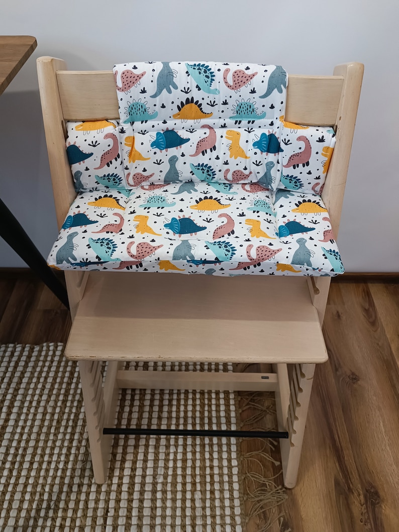 May include: A wooden high chair with a dinosaur-themed cushion set. The cushion set includes a back cushion, a seat cushion, and two side cushions. The cushions are white with a colourful dinosaur pattern.