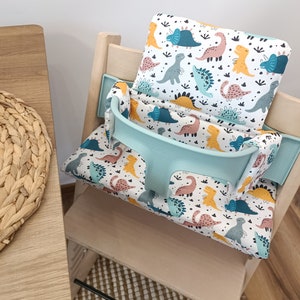 May include: A light blue and white high chair cushion with a dinosaur print. The cushion has a light blue strap and is placed on a wooden high chair.