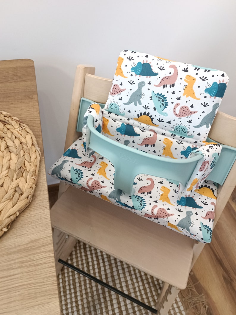 May include: A light blue and white high chair cushion with a dinosaur print. The cushion has a light blue strap and is placed on a wooden high chair.