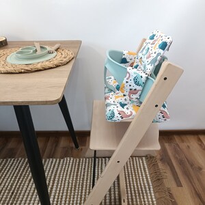 May include: A light wood high chair with a turquoise and white dinosaur patterned seat cushion. The chair is set up next to a wooden table with a woven placemat and a green silicone plate.
