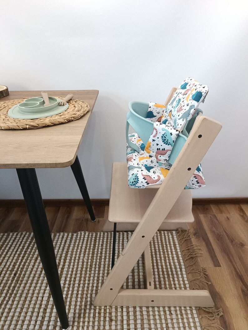 May include: A light wood high chair with a turquoise and white dinosaur patterned seat cushion. The chair is set up next to a wooden table with a woven placemat and a green silicone plate.
