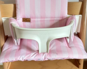 May include: A light wood high chair featuring a white plastic safety rail with the word STOKKE embossed on the front. The chair includes a padded seat and backrest cushion with a vertical light pink and white striped pattern and a small brown label.
