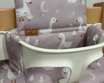 May include: A high chair cushion with a whimsical goose pattern in white and light brown on a lavender background. The cushion is designed to fit a Stokke high chair, providing comfort and style for the baby.