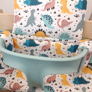 May include: A turquoise and white high chair cushion with a dinosaur print. The cushion has a white background with colourful dinosaurs in various poses. The text "STOKKE" is printed on the front of the cushion.
