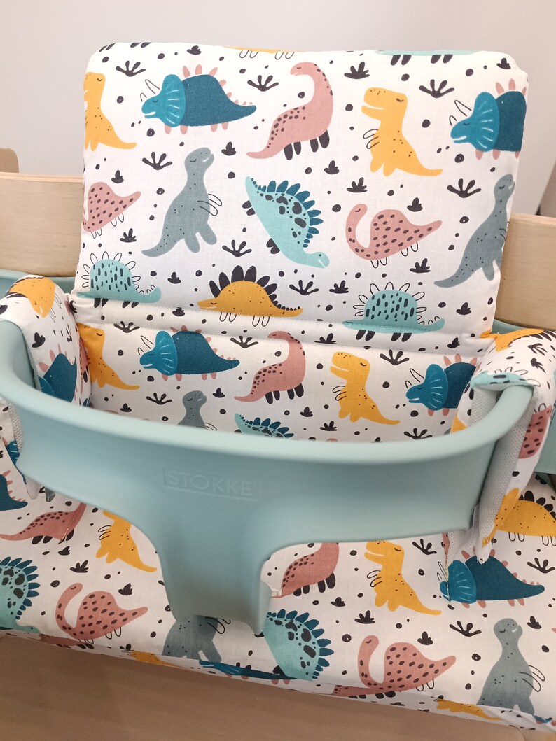 May include: A turquoise and white high chair cushion with a dinosaur print. The cushion has a white background with colourful dinosaurs in various poses. The text "STOKKE" is printed on the front of the cushion.