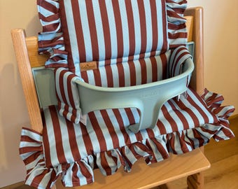 May include: A wooden high chair featuring a light green plastic safety rail and a padded fabric cushion set. The cushion has a vertical striped pattern in dark red and light blue with ruffled edges. The Stokke brand name is embossed on the rail.