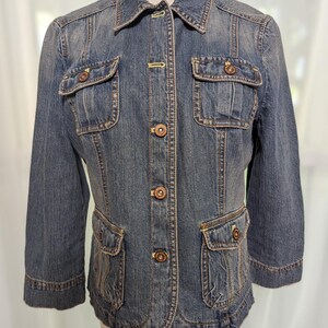 May include: A blue denim jacket with three-quarter sleeves. The jacket features four front pockets, a button-up closure, and decorative buttons. The jacket has a classic collar and a tailored fit, suitable for casual wear.