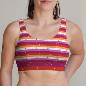 May include: A scoop-neck sports bra featuring horizontal stripes in red, orange, and purple, with white star accents. The bra has a racerback design and is made of a soft, stretchy material. The overall theme is pride and inclusivity.