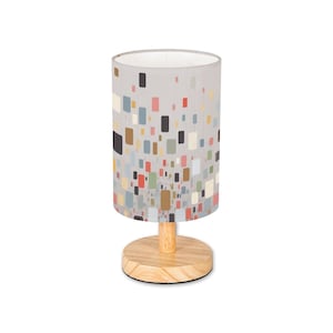 May include: A cylindrical table lamp with a light gray lampshade adorned with a pattern of colorful rectangles. The lamp has a wooden base and a wooden stem. The base is round and the stem is cylindrical.