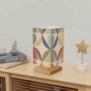 May include: A table lamp with a geometric patterned lampshade in shades of green, blue, yellow, and red. The lampshade is made of fabric and has a white base. The lamp is sitting on a wooden table.