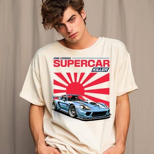 May include: Cream-colored t-shirt featuring a blue sports car graphic with a red sun and the text "JDM LEGEND SUPERCAR KILLER." The car has a sleek design with a spoiler and racing wheels.