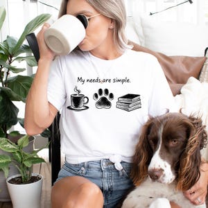 Op de afbeelding: Wit T-shirt met de tekst "My needs are simple" en illustraties van een koffiekopje, een pootafdruk en boeken. Een bruin-witte hond staat op de voorgrond. De persoon draagt een blauwe spijkerbroek en drinkt uit een witte mok.
