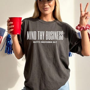 以下が含まれることがあります： 「MIND THY BUSINESS」と「PETTY PROVERBS 24:7」の白い文字が入った濃いグレーのTシャツ。赤いカップを持ち、ピースサインをしている。背景にはアメリカの国旗と装飾がある。
