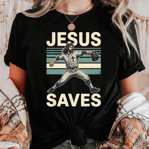 Puede incluir: Una camiseta negra de manga corta con un gráfico de un jugador de béisbol con pelo largo y barba, vistiendo un uniforme a rayas con una cruz en el pecho. El texto JESUS SAVES está impreso en letras grandes de color crema sobre el diseño.