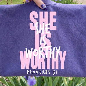 Puede incluir: Una sudadera morada con el texto "SHE IS WORTHY" en letras rosas y "PROVERBIOS 31" en blanco. La sudadera se muestra frente a un jardín con flores moradas y azules. El texto es en negrita.