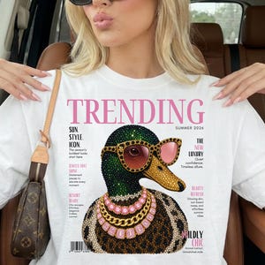 Może przedstawiać: Biały t-shirt z grafiką kaczki krzyżówki w różowych okularach przeciwsłonecznych oraz warstwowych naszyjnikach ze złota i różowych kamieni szlachetnych. Projekt imituje okładkę magazynu z napisem TRENDING, SUMMER 2026 oraz różnymi tekstami.
