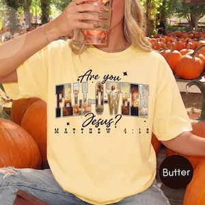 May include: A butter yellow t-shirt featuring the text Are you FALL-O-WEEN Jesus? and Matthew 4:19. The letters are filled with photographic imagery of pumpkins and autumn scenes. Small star icons are placed around the central graphic text.