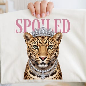 Op de afbeelding: Crèmekleurig T-shirt met een luipaard met een zilveren kroon en ketting. Het woord "SPOILED" is roze gedrukt boven de kop van de luipaard. De luipaard heeft een gevlekte vacht en groene ogen.