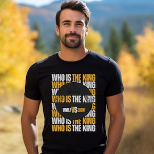 Puede incluir: Una camiseta negra con un estampado gráfico blanco que dice "WHO IS THE KING" varias veces. Un círculo negro con un gráfico de corona dorada y el texto "WOLF VS LION" está centrado en la camiseta.