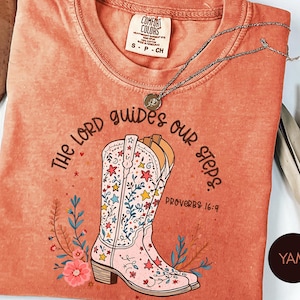 May include: A terracotta-colored t-shirt featuring a graphic of a light pink cowboy boot decorated with colorful stars and floral patterns. The text reads "THE LORD guides our steps." and "PROVERBS 16:9" in dark brown lettering. A silver necklace rests on the shirt.