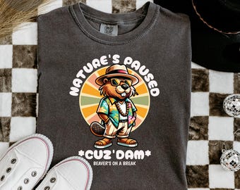 Puede incluir: Una camiseta gris oscuro con un castor de dibujos animados que lleva gafas de sol y una camisa hawaiana. El texto "Nature's Paused *Cuz'Dam* Beaver's on a Break" está impreso en la camiseta.