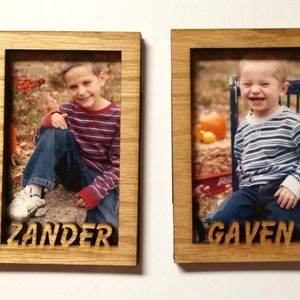 May include: Two wooden picture frames with a light brown wood finish. Each frame has a photo of a young boy. The frame on the left has the name "Zander" engraved below the photo. The frame on the right has the name "Gaven" engraved below the photo.