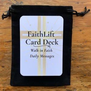 May include: A black velvet drawstring bag containing a deck of cards. The cards are white with gold accents and the text "FaithLift Card Deck", "Walk in Faith", and "Daily Messages". The bag is on a wooden surface.