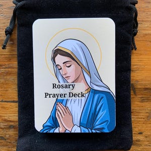 May include: A black velvet drawstring bag holds a deck of cards. The cards feature an illustration of a woman in a blue veil with a gold halo. The text "Rosary Prayer Deck" is printed on the card.