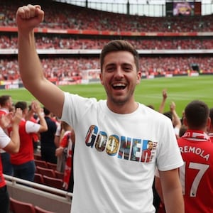 Può includere: Maglietta bianca con la scritta "GOONER" a lettere colorate. La persona nell'immagine sorride e alza il pugno in aria. Lo sfondo mostra uno stadio pieno di persone.