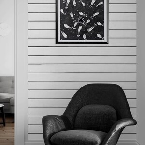 May include: A black and white framed artwork featuring a pattern of ghost-like figures on a dark background hangs above a black armchair. The wall is white with horizontal black stripes. The room has wooden flooring.