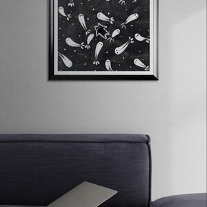May include: Black framed art print featuring a whimsical illustration of white ghost-like figures on a dark background with white dots. A black cat-like figure is in the center. The artwork is displayed on a wall above a dark blue couch with a laptop.