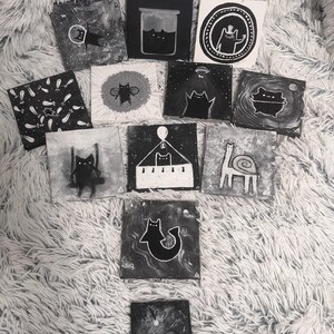 May include: A collection of small, square, black and white paintings featuring whimsical cat illustrations. The artwork includes cats in various scenarios, such as in space, in a jar, and on a swing. The paintings are displayed on a fluffy, white surface.