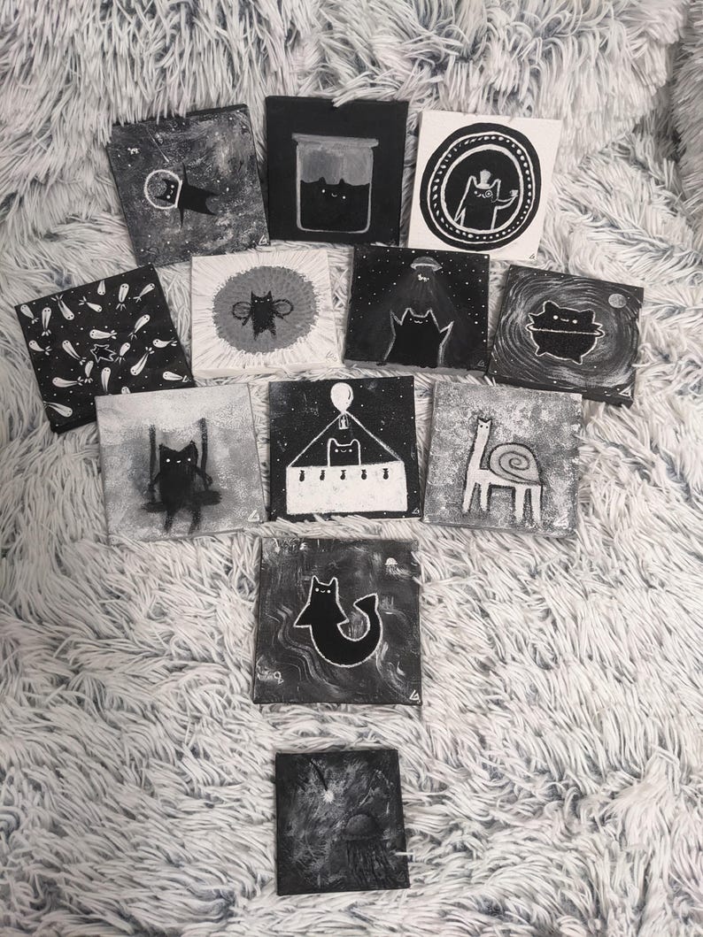 May include: A collection of small, square, black and white paintings featuring whimsical cat illustrations. The artwork includes cats in various scenarios, such as in space, in a jar, and on a swing. The paintings are displayed on a fluffy, white surface.