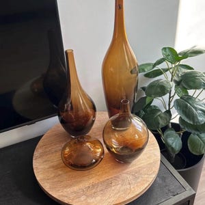 Op de afbeelding: Een verzameling amberkleurige glazen vazen in verschillende vormen en maten. De vazen staan op een ronde houten schaal. De hoogste vaas is slank en elegant. De opstelling staat tegen een neutrale achtergrond, met een potplant. De vazen zijn van glas.