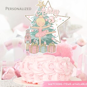 May include: A pink and green Christmas-themed cake topper featuring a ballerina, a nutcracker, a Christmas tree, and presents. The topper is personalized with the name "Ashley" and the number "3".
