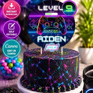 以下が含まれることがあります： カラフルな飛沫模様の黒い丸いケーキ。ゲームをテーマにしたケーキトッパーが飾られています。トッパーにはゲームコントローラー、「LEVEL 9」、「GAME ON AIDEN」、「LEVEL UP」の文字が表示されています。ケーキは白いケーキスタンドの上にあります。