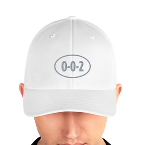 以下が含まれることがあります： 「0-0-2」と書かれたグレーの楕円形のワッペンが付いた白いベースボールキャップ。