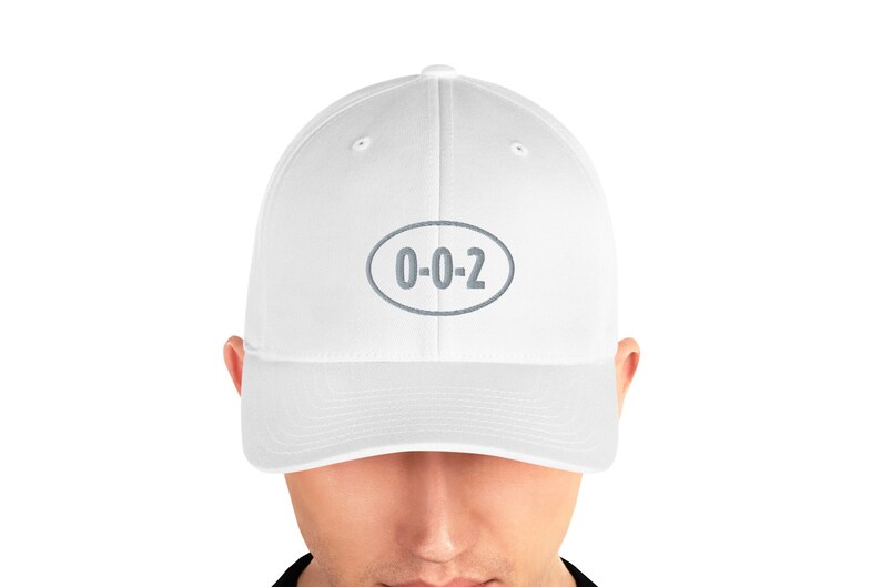 以下が含まれることがあります： 「0-0-2」と書かれたグレーの楕円形のワッペンが付いた白いベースボールキャップ。
