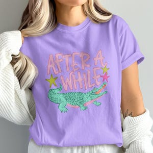 May include: A lilac-colored t-shirt featuring a cartoon alligator with the text "AFTER A WHILE" in pink and yellow. The alligator is green with pink accents and is surrounded by yellow and pink stars.
