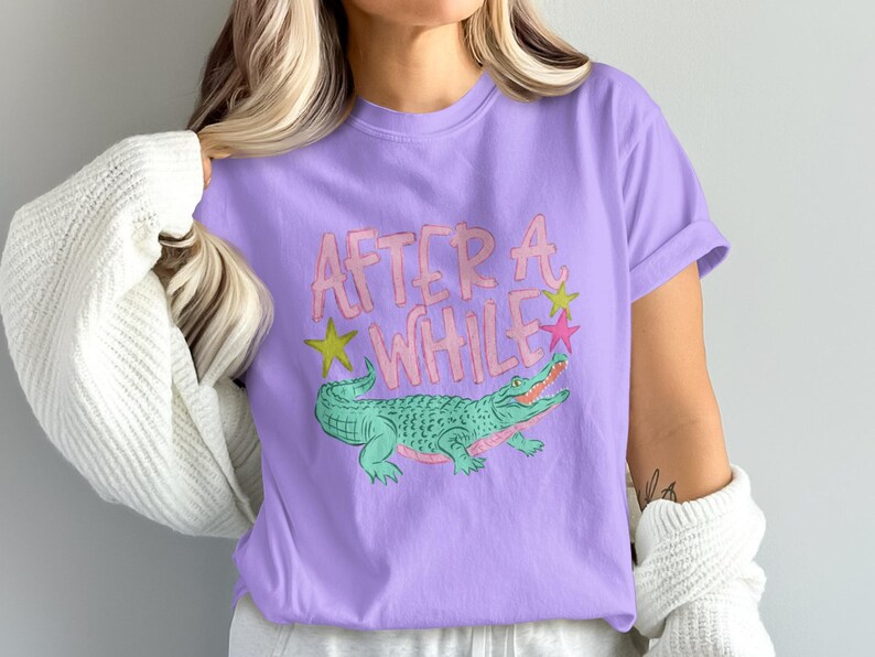 May include: A lilac-colored t-shirt featuring a cartoon alligator with the text "AFTER A WHILE" in pink and yellow. The alligator is green with pink accents and is surrounded by yellow and pink stars.